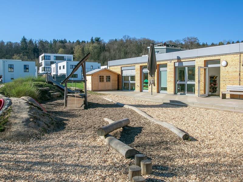 Ein moderner Spielplatz mit Rutsche, Klettergerüst und Holzhütte befindet sich vor einem flachen, einstöckigen Gebäude, umgeben von Bäumen und mehrstöckigen weißen Wohnhäusern.