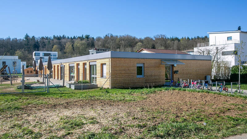 Ein einstöckiges Gebäude mit Holzfassade steht in einer grünen Umgebung, flankiert von Fahrrädern und umgeben von Wohnhäusern im Hintergrund.