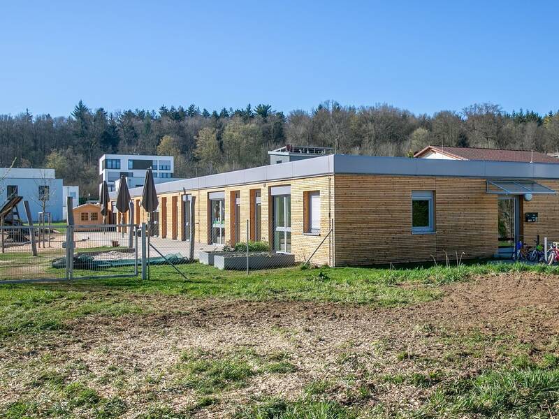 Ein einstöckiges Gebäude mit Holzfassade steht in einer grünen Umgebung, flankiert von Fahrrädern und umgeben von Wohnhäusern im Hintergrund.