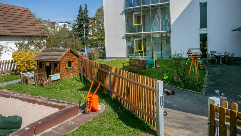 Ein kleiner Spielplatz mit einem Holzhäuschen, einem Sandkasten und verschiedenen Spielgeräten befindet sich vor einem modernen Gebäude mit großen Fenstern.