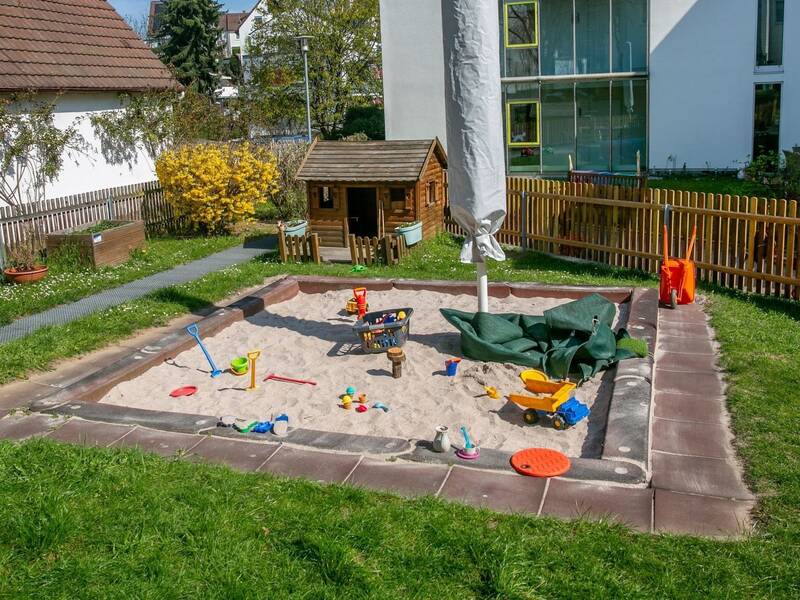 Ein Sandkasten mit verschiedenen bunten Spielzeugen, umgeben von einem Garten mit Gras und Zäunen, steht neben einem kleinen Holzhaus und einem geschlossenen Sonnenschirm.