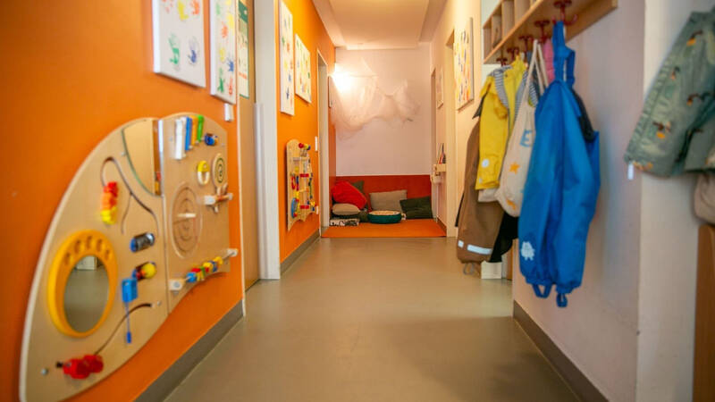 Ein farbenfroher Kindergartenflur mit Spielzeugwänden und an Haken hängender Kleidung führt zu einem gemütlichen Sitzbereich mit Kissen.