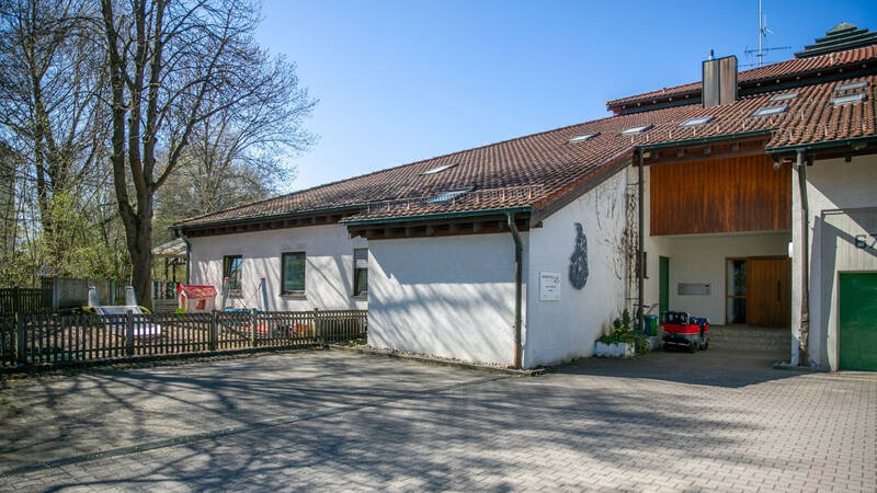 Ein Gebäude mit Satteldach und weißen Wänden steht neben einem eingezäunten Spielplatz mit Spielgeräten, umgeben von Bäumen.