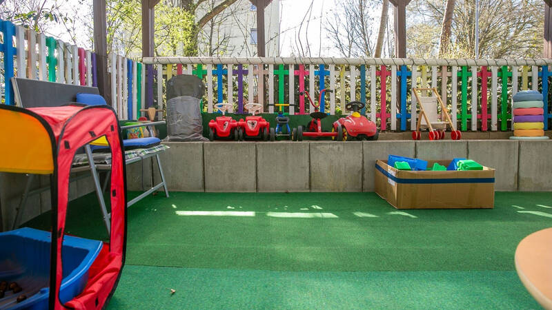 Ein Spielbereich im Freien mit bunten Zäunen zeigt Spielzeugfahrzeuge, einen roten Spieltunnel, eine Wippe und eine Kiste mit Sandschaufeln.