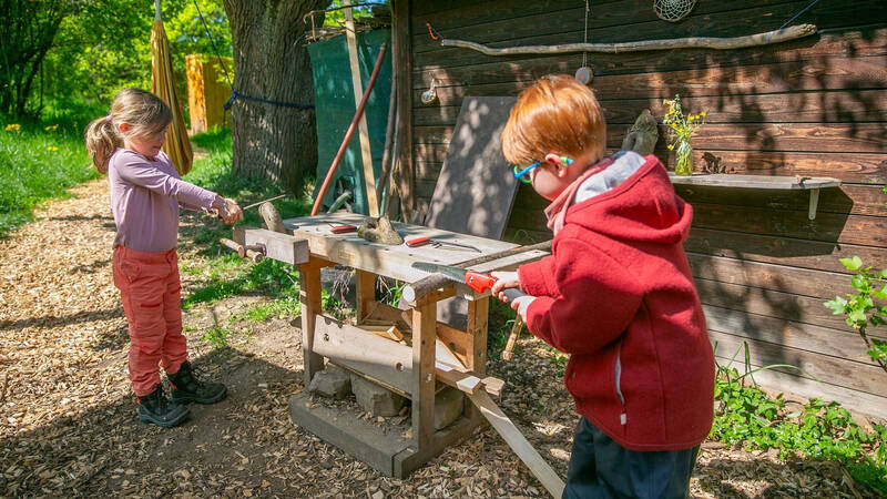 Zwei Kinder arbeiten im Freien an einem Holztisch mit Werkzeugen, umgeben von Bäumen und einem hölzernen Gebäude.