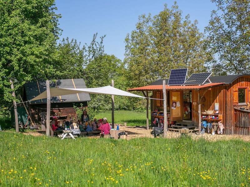 Vor einem kleinen Holzhaus mit Solarpanels, umgeben von Bäumen und grünem Gras, sitzen mehrere Kinder unter einem weißen Sonnensegel.