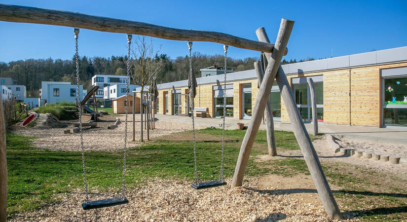 Der Spielplatz mit Schaukeln und Klettergeräten befindet sich vor einem modernen, holzverkleideten Gebäude mit großen Fenstern.