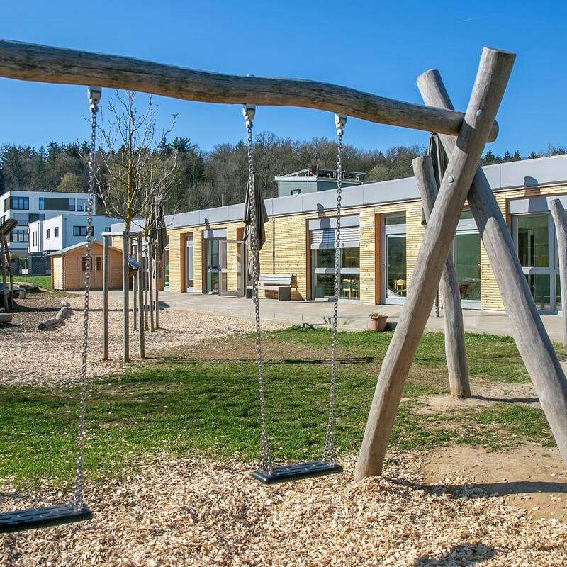 Der Spielplatz mit Schaukeln und Klettergeräten befindet sich vor einem modernen, holzverkleideten Gebäude mit großen Fenstern.