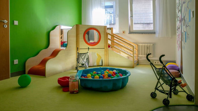 Ein bunter Kinderraum mit grünen Wänden und einer kleinen Rutsche, einem Bällebad, einem Kinderwagen und verschiedenen Spielsachen.