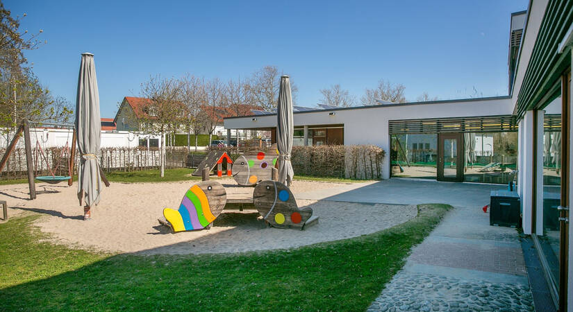 Ein moderner Spielplatz mit bunten Klettergeräten und Sonnenschirmen befindet sich in einem offenen Außenbereich neben einem Gebäude.
