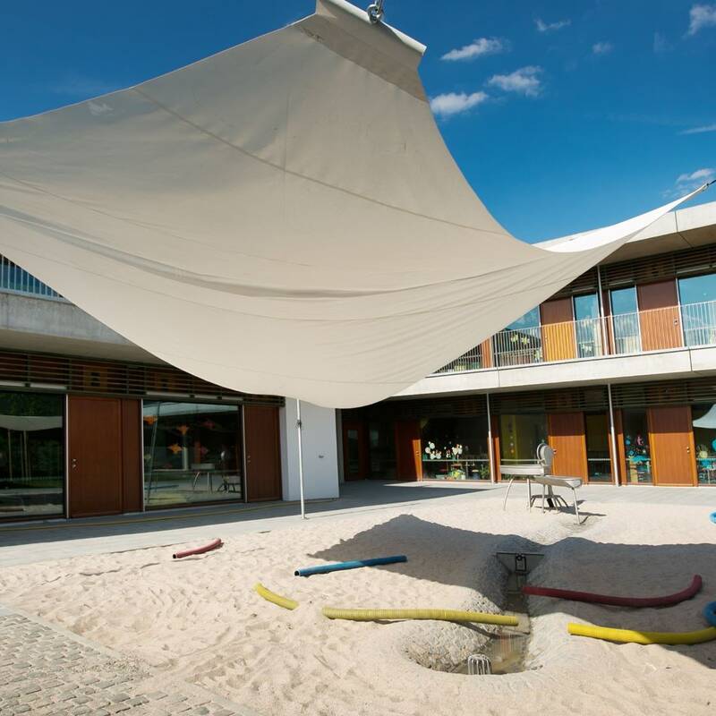 Großes Sonnensegel über einem Sandspielplatz vor einem großen Gebäude aus Holz